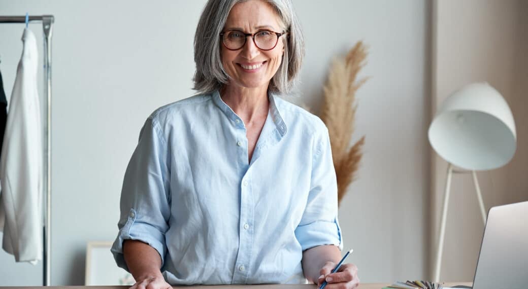 Happy middle aged stylish woman fashion designer wears glasses standing at workplace desk, portrait. Smiling sophisticated 60s old lady dressmaker drawing sketches, looking at camera in modern atelier