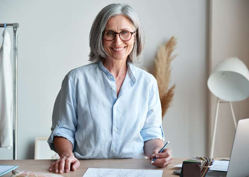 Happy middle aged stylish woman fashion designer wears glasses standing at workplace desk, portrait. Smiling sophisticated 60s old lady dressmaker drawing sketches, looking at camera in modern atelier