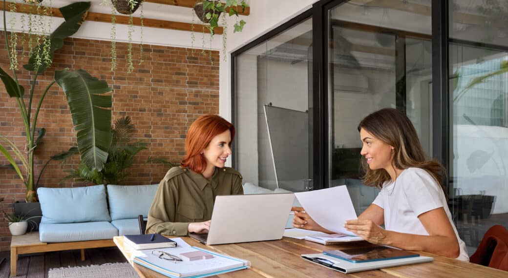 Two happy busy business women talking working together in green cozy office sitting at desk. Professional manager consulting client having conversation holding documents using laptop at work.