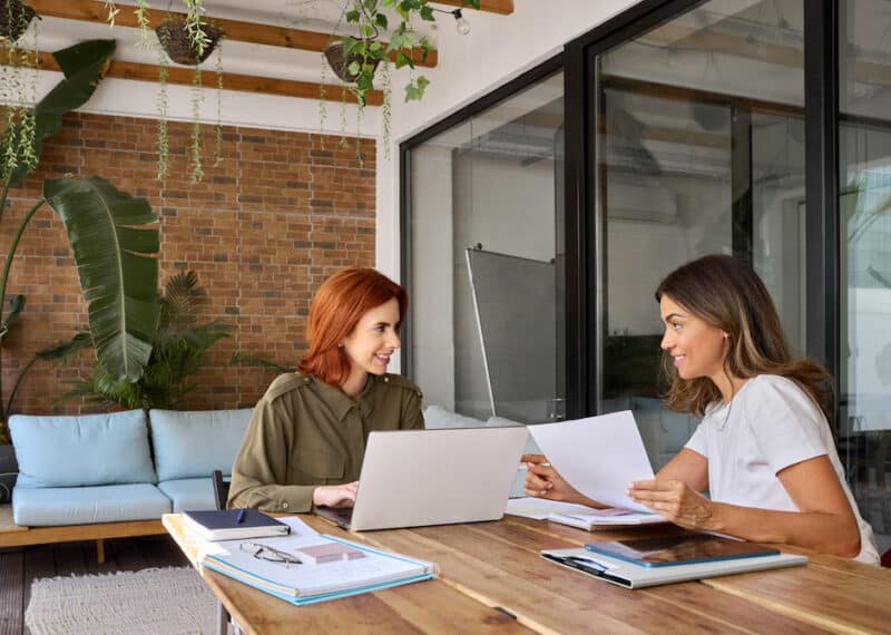 Two happy busy business women talking working together in green cozy office sitting at desk. Professional manager consulting client having conversation holding documents using laptop at work.