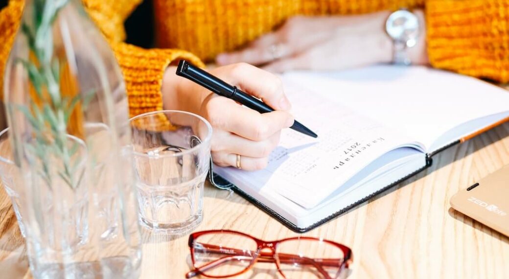 Glasses and vase sit on a wooden desk while woman in orange sweater writes with a pen in a planner.