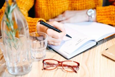 Glasses and vase sit on a wooden desk while woman in orange sweater writes with a pen in a planner.