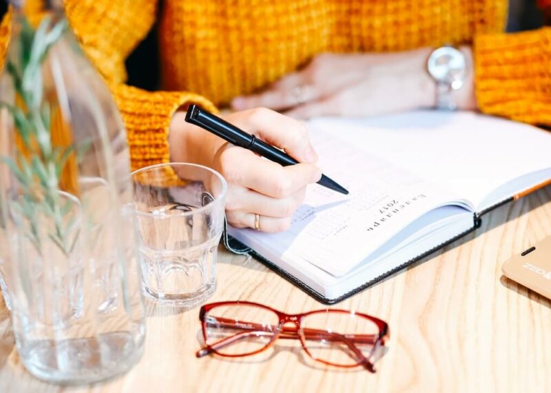 Glasses and vase sit on a wooden desk while woman in orange sweater writes with a pen in a planner.