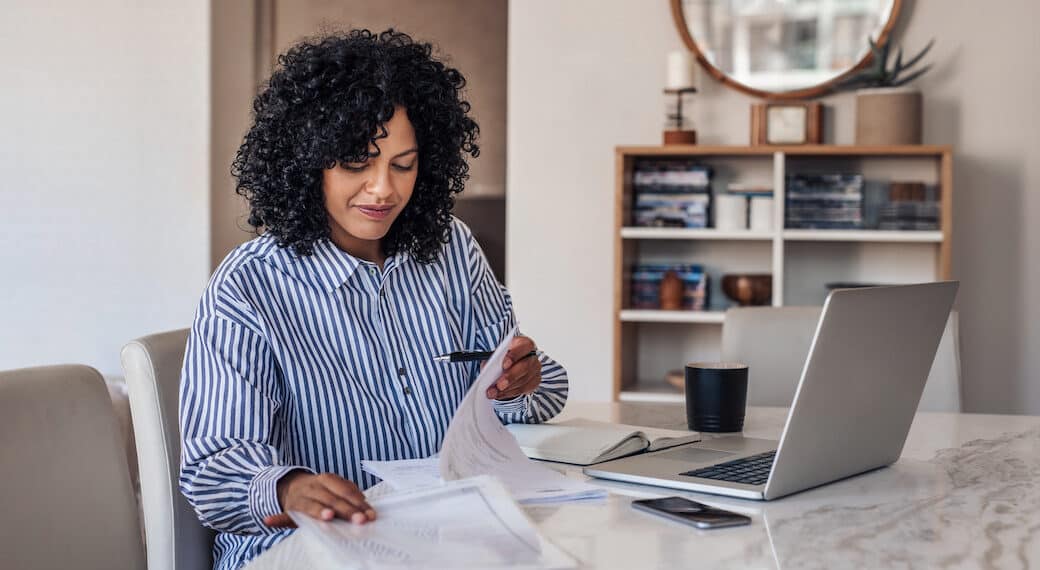 Smiling young female entrepreneur going through paperwork while working on a laptop at her dining room table at home