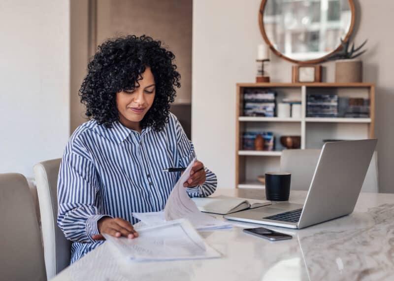 Smiling young female entrepreneur going through paperwork while working on a laptop at her dining room table at home