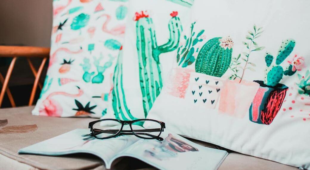 Three pillows with bright and colorful cacti illustrations printed on them on a tan couch with a magazine and glasses