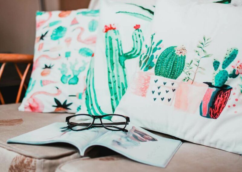 Three pillows with bright and colorful cacti illustrations printed on them on a tan couch with a magazine and glasses