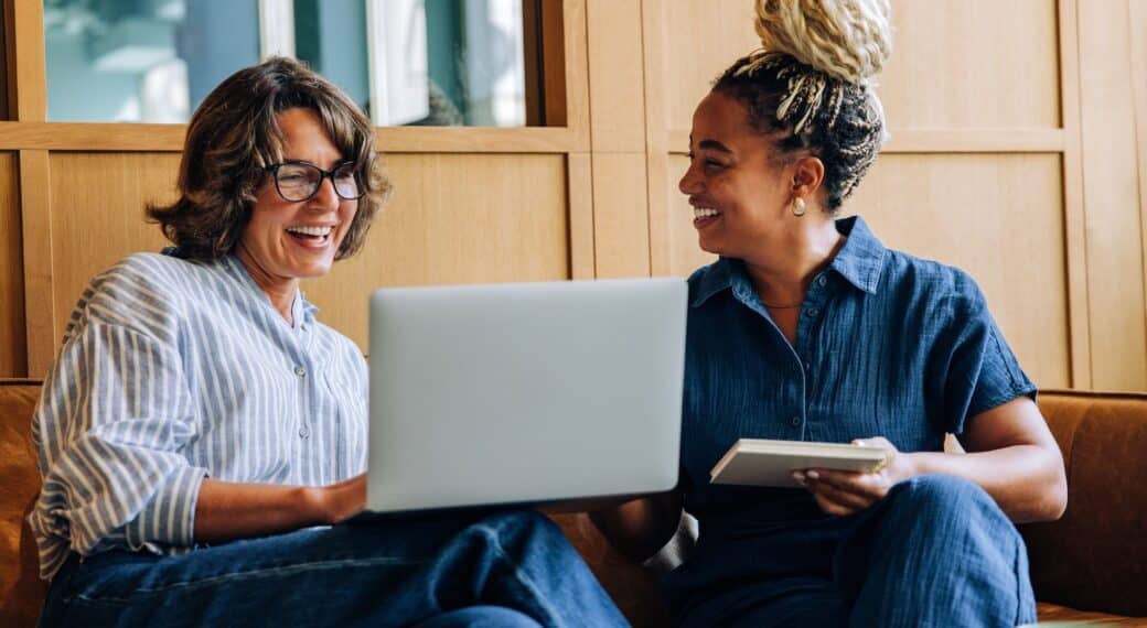 Two adult women enjoy a friendly discussion, using a laptop and a notebook. Their bright smiles convey positivity, teamwork, and productivity in a modern work environment.