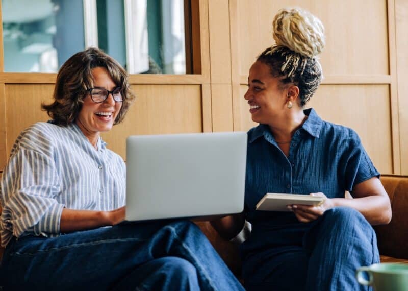 Two adult women enjoy a friendly discussion, using a laptop and a notebook. Their bright smiles convey positivity, teamwork, and productivity in a modern work environment.